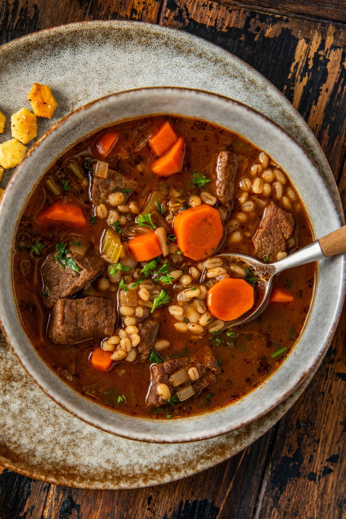 Made easy in one-pot, Beef Barley Soup is loaded with tender beef, chewy pearl barley, and lots of vegetables in a rich and savory broth. A bowl of beef barley soup with chunks of beef, sliced carrots, barley, and herbs in a rich broth, served on a rustic plate with oyster crackers on the side. A spoon rests in the bowl.
