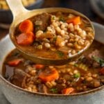 A ladle of beef barley soup with pieces of beef, barley, carrots, and herbs being served into a ceramic bowl.
