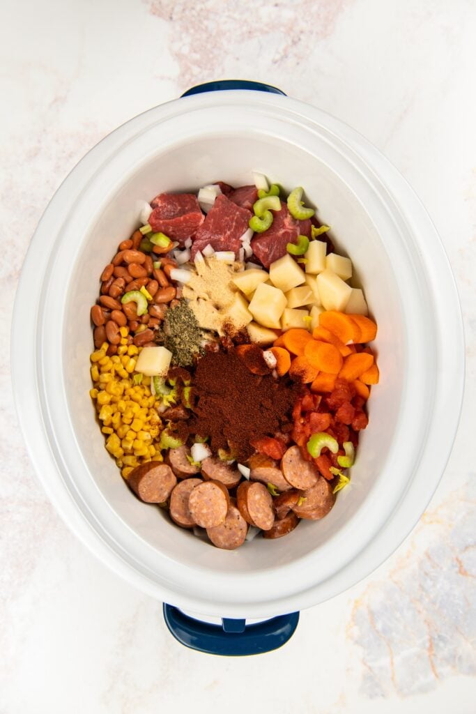 This hearty Crockpot Cowboy Stew is loaded with tender beef, sausage, beans, and veggies for the ultimate one-pot comfort food dinner. A slow cooker filled with raw ingredients arranged in sections: chopped beef, sliced sausage, celery, potatoes, carrots, corn, beans, chopped onions, and various seasonings on top.