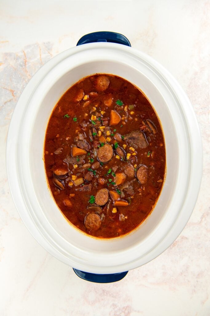 This hearty Crockpot Cowboy Stew is loaded with tender beef, sausage, beans, and veggies for the ultimate one-pot comfort food dinner. A white slow cooker filled with a rich, chunky stew featuring sliced sausage, beans, corn, and vegetables in a dark broth, garnished with chopped herbs.