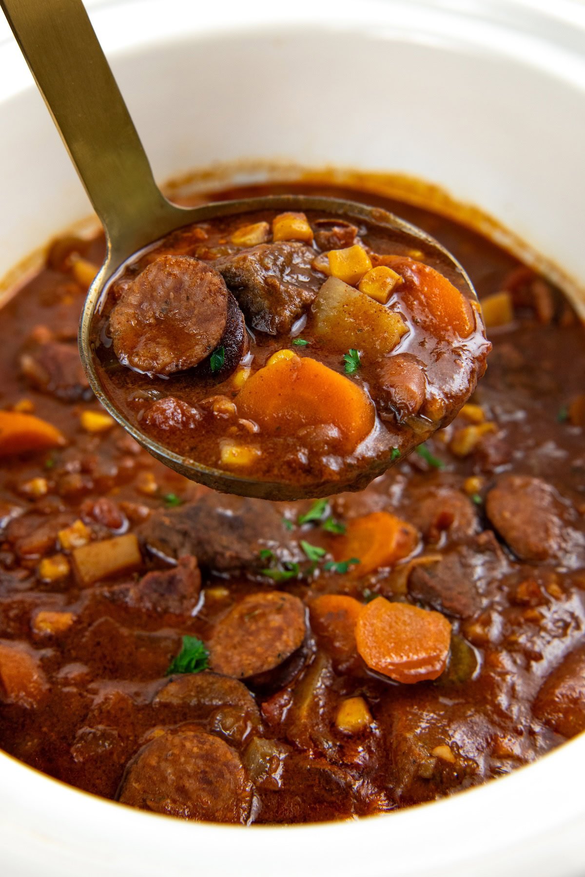 This hearty Crockpot Cowboy Stew is loaded with tender beef, sausage, beans, and veggies for the ultimate one-pot comfort food dinner. A close-up of a ladle lifting cowboy stew from a crockpot, showing sliced sausage, carrots, potatoes, and corn in a rich, reddish broth.