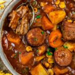 A bowl of crockpot cowboy stew featuring chunks of beef, sausage, potatoes, carrots, and corn in a rich, brown broth. A spoon rests in the bowl, lifting a piece of beef. A slice of buttered cornbread is nearby.