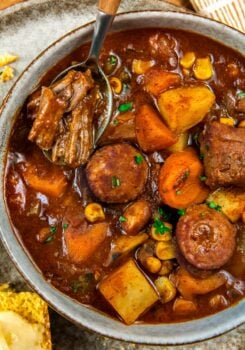 A bowl of crockpot cowboy stew featuring chunks of beef, sausage, potatoes, carrots, and corn in a rich, brown broth. A spoon rests in the bowl, lifting a piece of beef. A slice of buttered cornbread is nearby.