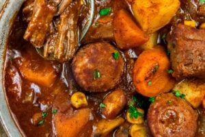 A bowl of crockpot cowboy stew featuring chunks of beef, sausage, potatoes, carrots, and corn in a rich, brown broth. A spoon rests in the bowl, lifting a piece of beef. A slice of buttered cornbread is nearby.