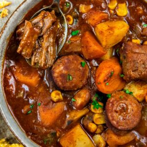 A bowl of crockpot cowboy stew featuring chunks of beef, sausage, potatoes, carrots, and corn in a rich, brown broth. A spoon rests in the bowl, lifting a piece of beef. A slice of buttered cornbread is nearby.