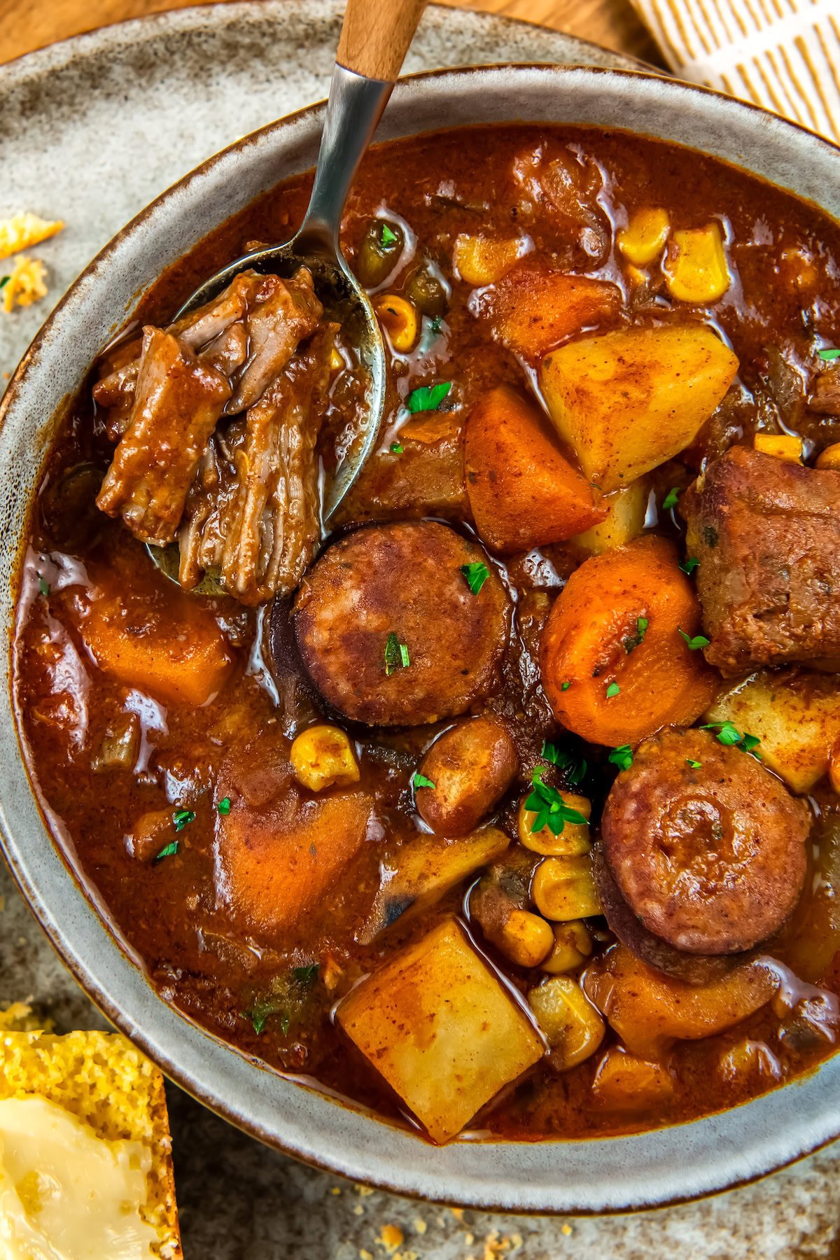 This hearty Crockpot Cowboy Stew is loaded with tender beef, sausage, beans, and veggies for the ultimate one-pot comfort food dinner. A bowl of crockpot cowboy stew featuring chunks of beef, sausage, potatoes, carrots, and corn in a rich, brown broth. A spoon rests in the bowl, lifting a piece of beef. A slice of buttered cornbread is nearby.
