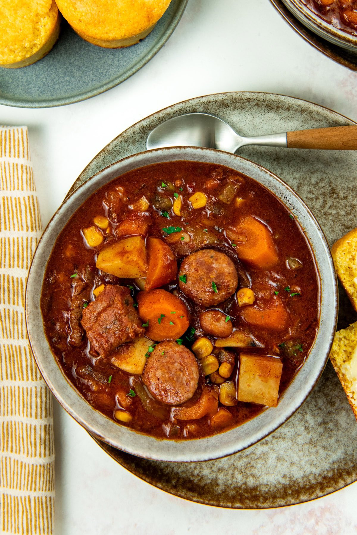 This hearty Crockpot Cowboy Stew is loaded with tender beef, sausage, beans, and veggies for the ultimate one-pot comfort food dinner. A bowl of crockpot cowboy stew with sausage slices, beef chunks, potatoes, carrots, and chickpeas in a rich broth, served on a plate with a spoon. Cornbread muffins and a striped napkin are nearby.