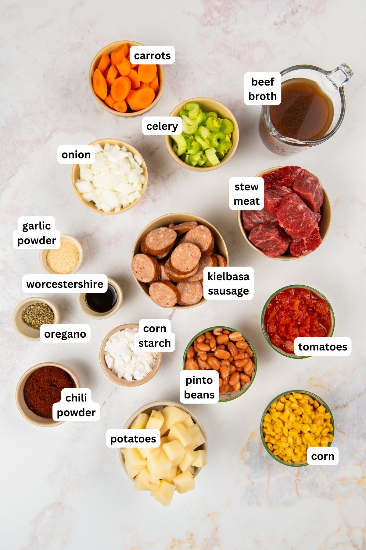 This hearty Crockpot Cowboy Stew is loaded with tender beef, sausage, beans, and veggies for the ultimate one-pot comfort food dinner. Labeled ingredients in bowls for crockpot cowboy stew recipe: sliced carrots, celery, chopped onion, garlic powder, Worcestershire sauce, oregano, chili powder, potatoes, beef broth, stew meat, kielbasa sausage, corn starch, pinto beans, tomatoes, and corn.