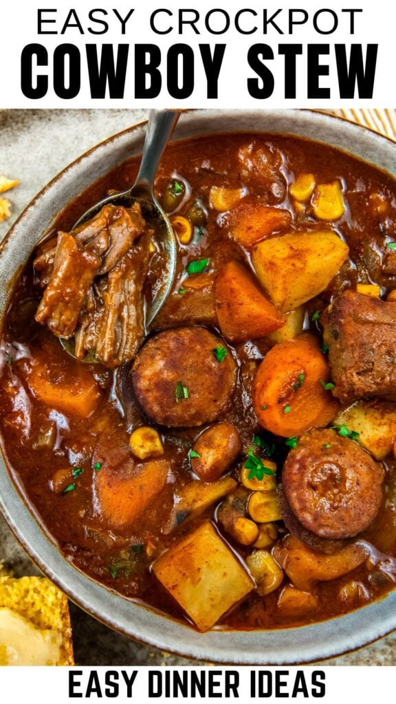 A bowl of hearty cowboy stew with chunks of beef, sausage slices, potatoes, carrots, corn, and beans in a rich broth, garnished with parsley. A spoon holds a piece of meat above the bowl of beef stew.