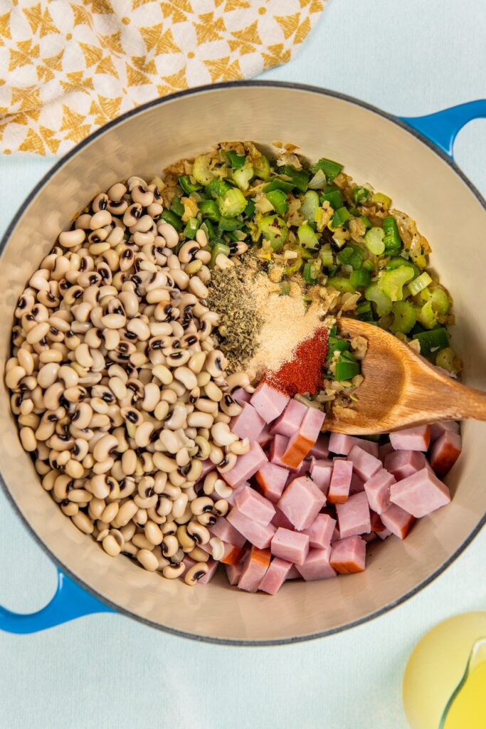 Slowly simmered in a rich and smoky broth, these Southern Black Eyed Peas are insanely flavorful and tender as can be! A blue pot contains black-eyed peas, chopped ham, diced celery and green pepper, and assorted spices, with a wooden spoon resting inside.