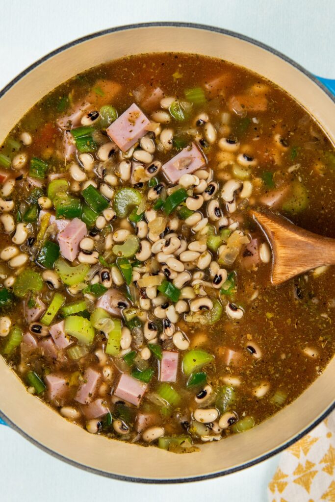 Slowly simmered in a rich and smoky broth, these Southern Black Eyed Peas are insanely flavorful and tender as can be! Peas, diced ham, celery, green onions, and more, in a large pot with a wooden spoon stirring the broth.