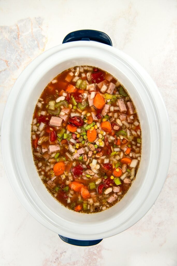 A white slow cooker filled with a colorful bean and vegetable soup containing carrots, celery, corn, tomatoes, green beans, and chunks of ham in a broth, viewed from above.