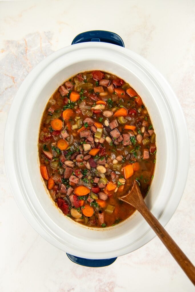 Crockpot 15 bean soup in a slow cooker, containing sliced carrots, beans, herbs, and diced ham, with a wooden spoon resting inside.