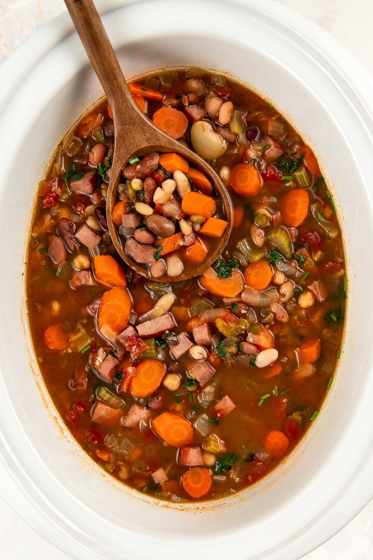 A white slow cooker filled with 15 bean soup with ham and vegetables with a wooden spoon scooping up a portion.