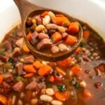 A wooden spoon lifts a serving of hearty 15 bean soup, showing chunks of carrots, beans, and pieces of ham in a rich broth, with the rest of the soup in a crockpot below.