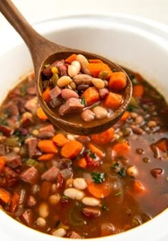A wooden spoon lifts a serving of hearty 15 bean soup, showing chunks of carrots, beans, and pieces of ham in a rich broth, with the rest of the soup in a crockpot below.