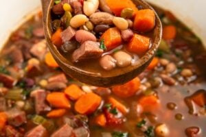 A wooden spoon lifts a serving of hearty 15 bean soup, showing chunks of carrots, beans, and pieces of ham in a rich broth, with the rest of the soup in a crockpot below.