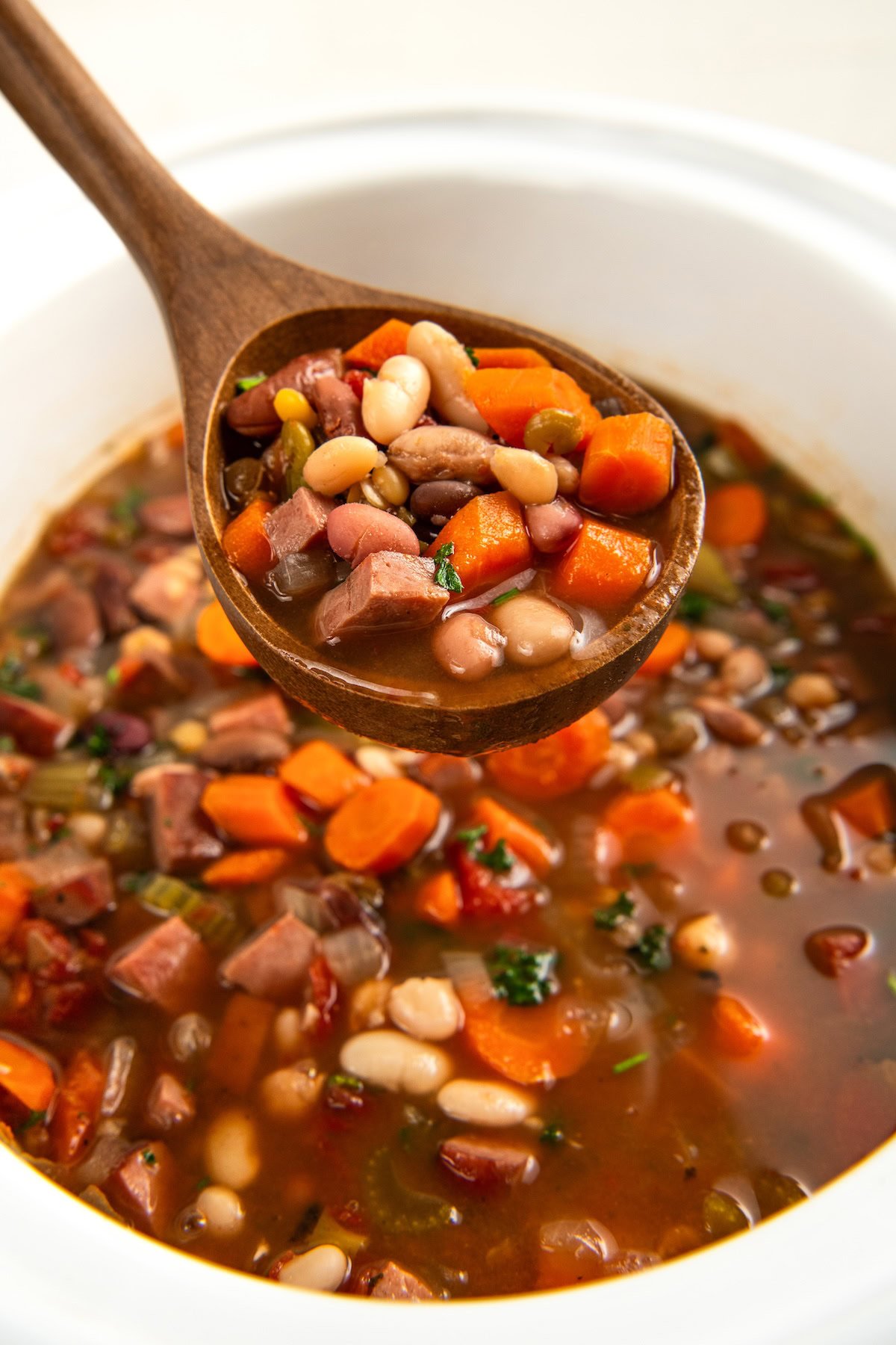A wooden spoon lifts a serving of hearty 15 bean soup, showing chunks of carrots, beans, and pieces of ham in a rich broth, with the rest of the soup in a crockpot below.