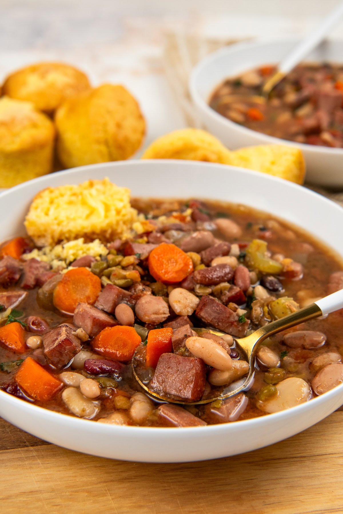 A bowl of 15 bean soup with ham, carrots, and celery, served with a spoon and a side of cornbread.