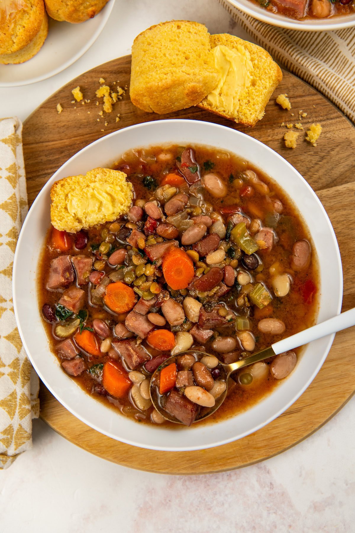 A bowl of hearty 15 bean soup with ham served on a wooden board with a spoon and a piece of buttered cornbread on the side.