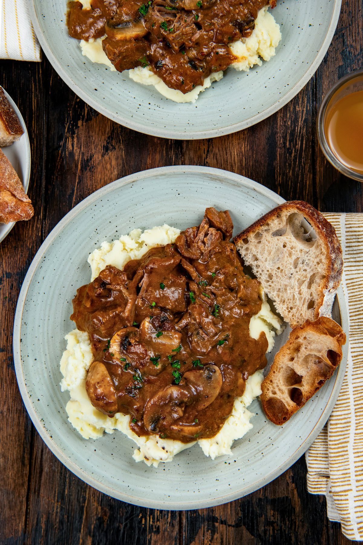 This easy Crockpot Steak Diane recipe slowly simmers ultra-tender beef and hearty mushrooms in a rich, creamy, and savory sauce. A plate of mashed potatoes topped with crockpot Steak Diane in a brown mushroom gravy, garnished with fresh herbs, served with two slices of rustic bread on the side.