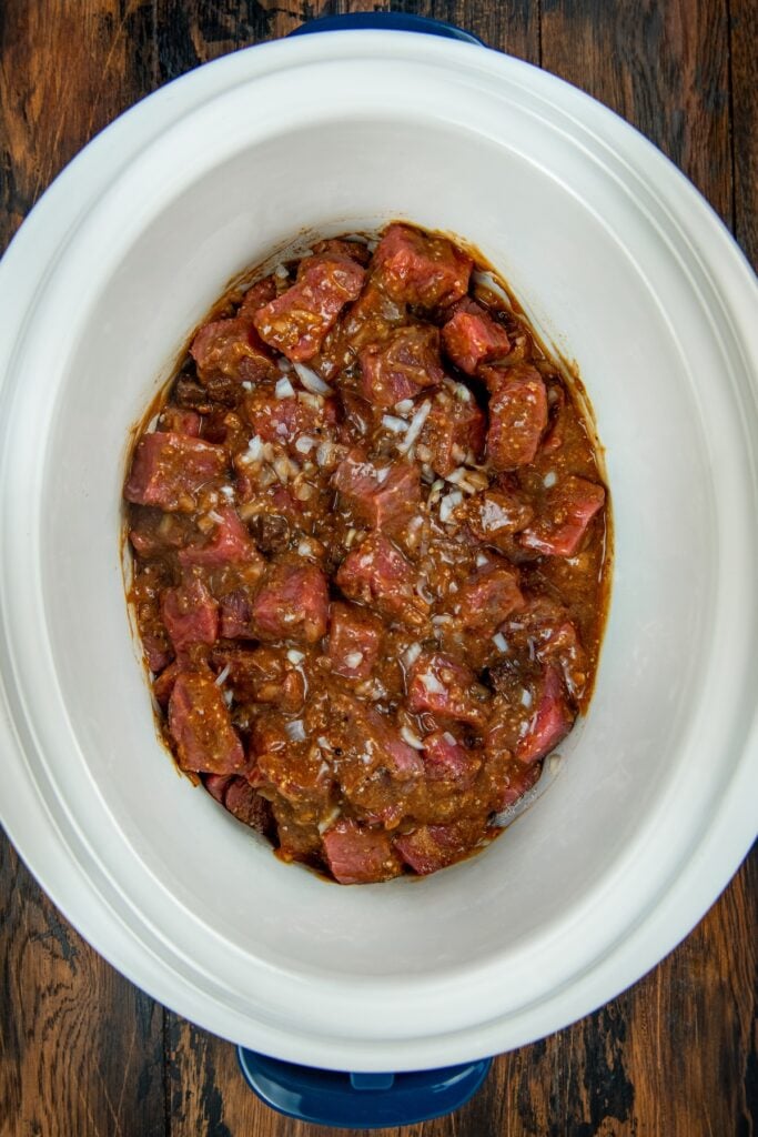 This easy Crockpot Steak Diane recipe slowly simmers ultra-tender beef and hearty mushrooms in a rich, creamy, and savory sauce. Chunks of raw beef stew meat marinated with sauce and chopped onions are placed inside a white slow cooker, ready for cooking.