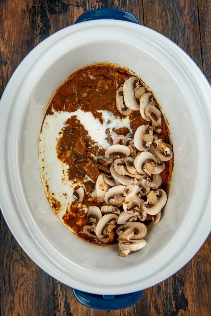 This easy Crockpot Steak Diane recipe slowly simmers ultra-tender beef and hearty mushrooms in a rich, creamy, and savory sauce. Cooked beef stew with brown sauce in a slow cooker topped with sliced raw mushrooms and cream.