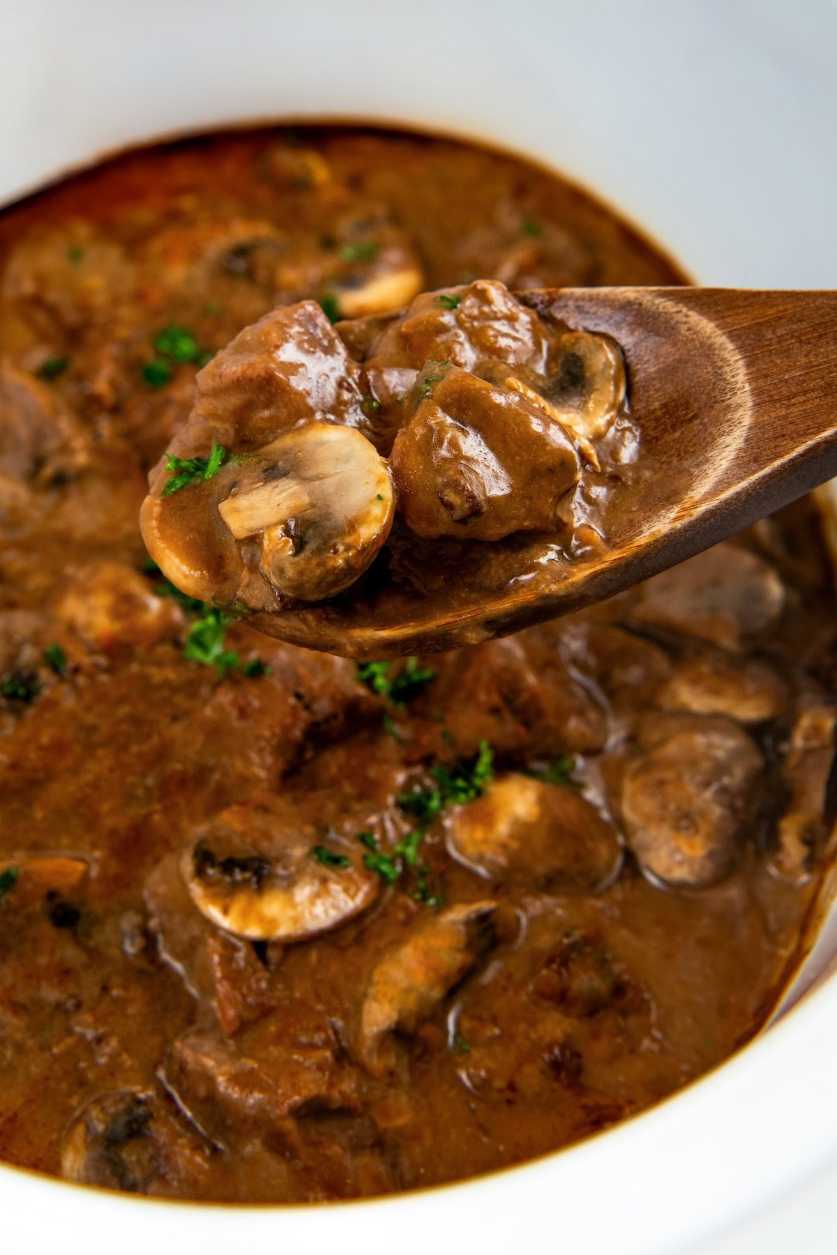 This easy Crockpot Steak Diane recipe slowly simmers ultra-tender beef and hearty mushrooms in a rich, creamy, and savory sauce. A wooden serving spoon is lifting crockpot steak Diane out of a slow cooker.