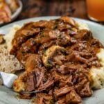 A plate of mashed potatoes topped with tender slow cooker steak Diane with rich brown gravy and mushrooms with fresh herbs on top.
