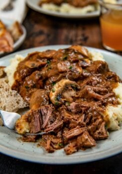 A plate of mashed potatoes topped with tender slow cooker steak Diane with rich brown gravy and mushrooms with fresh herbs on top.