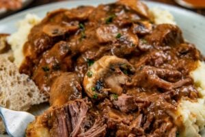 A plate of mashed potatoes topped with tender slow cooker steak Diane with rich brown gravy and mushrooms with fresh herbs on top.