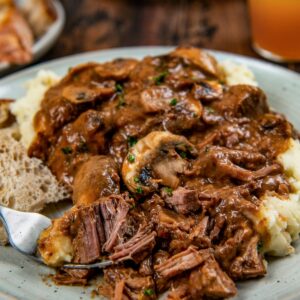A plate of mashed potatoes topped with tender slow cooker steak Diane with rich brown gravy and mushrooms with fresh herbs on top.