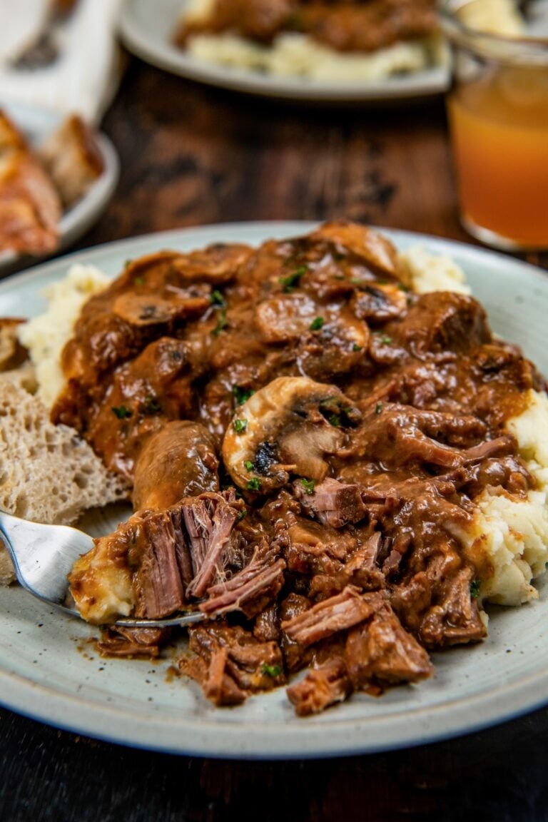 A plate of mashed potatoes topped with tender slow cooker steak Diane with rich brown gravy and mushrooms with fresh herbs on top.