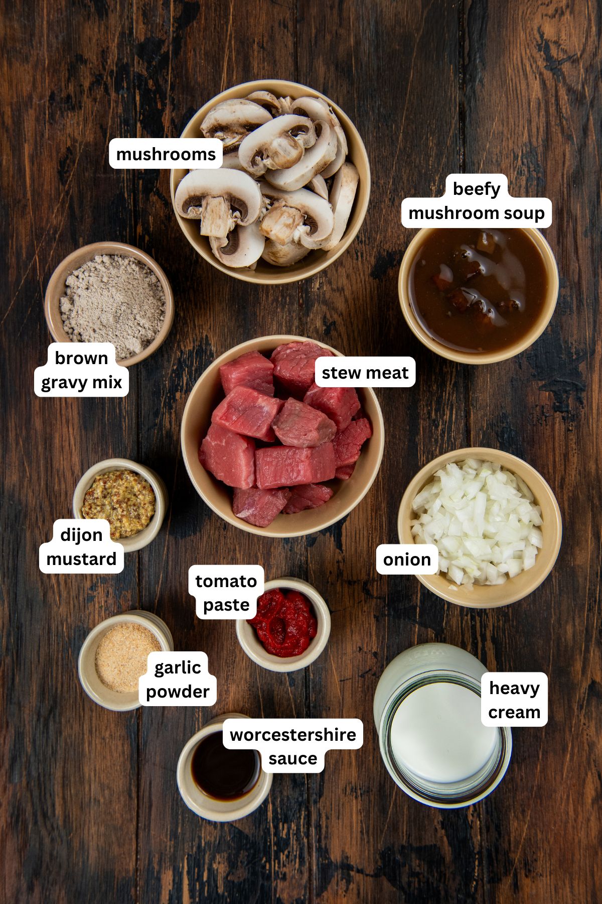 This easy Crockpot Steak Diane recipe slowly simmers ultra-tender beef and hearty mushrooms in a rich, creamy, and savory sauce. Labeled bowls of ingredients for crockpot steak Diane recipe; from top to bottom: sliced mushrooms, beefy mushroom soup, brown gravy mix, stew meat, chopped onion, heavy cream, dijon mustard, tomato paste, garlic powder, and Worcestershire sauce.