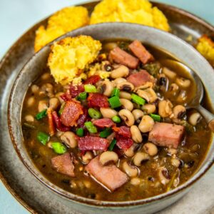 A bowl of southern black eyed peas with chunks of ham, topped with crispy bacon and chopped green onions, and served with two pieces of cornbread on a gray plate.