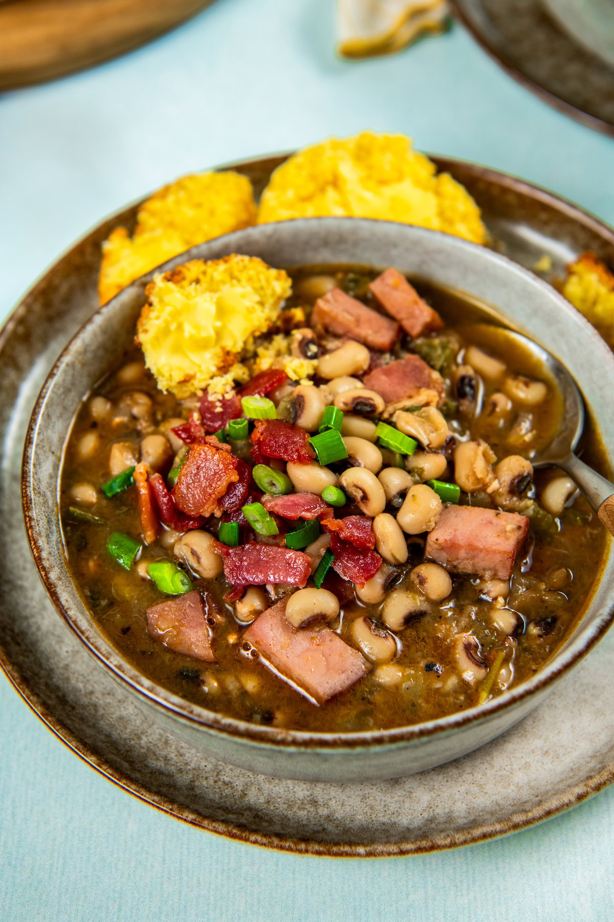 A bowl of southern black eyed peas with chunks of ham, topped with crispy bacon and chopped green onions, and served with two pieces of cornbread on a gray plate.