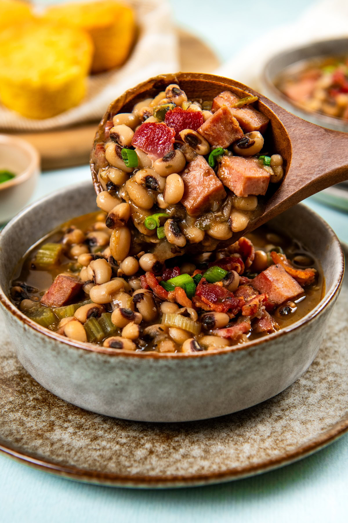 Slowly simmered in a rich and smoky broth, these Southern Black Eyed Peas are insanely flavorful and tender as can be! A wooden spoon pours a hearty serving of black eyed peas into a bowl with cornbread on the side of the plate.