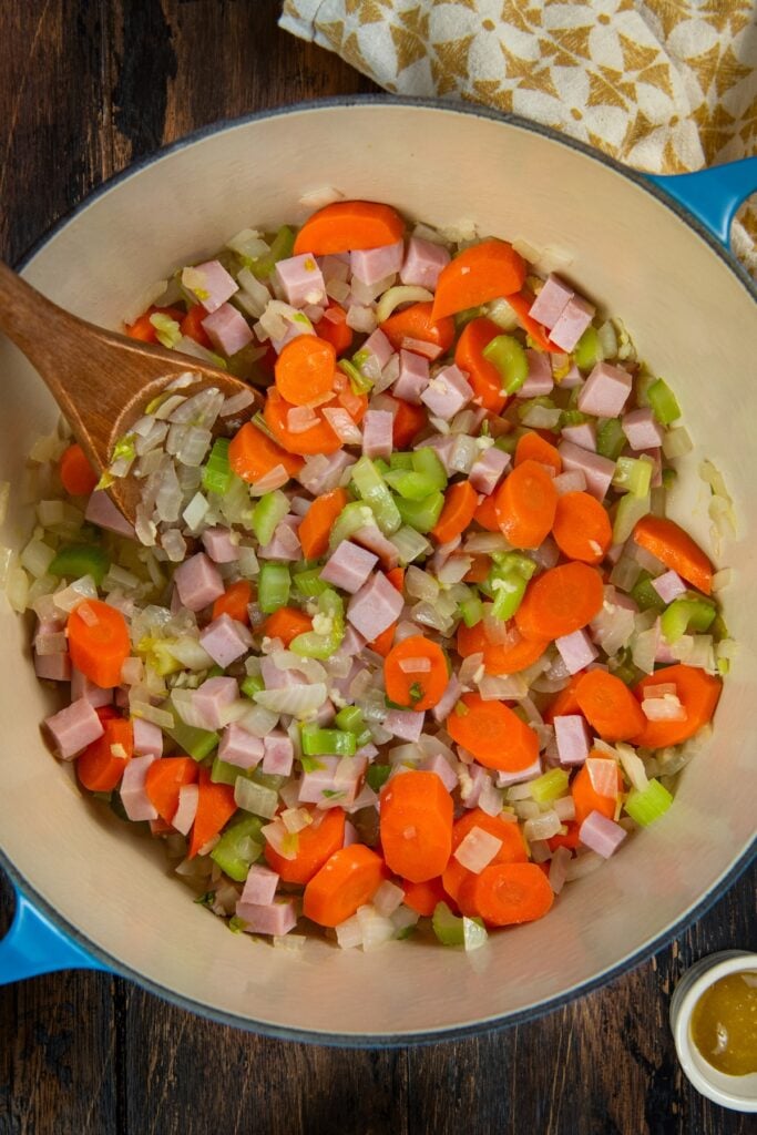 Made with dried or canned beans, this hearty Navy Bean Soup recipe is packed with creamy white beans, smoky ham, and flavorful veggies. A blue pot filled with chopped onions, celery, carrots, and diced ham being sautéed, stirred with a wooden spoon on a dark wooden surface.