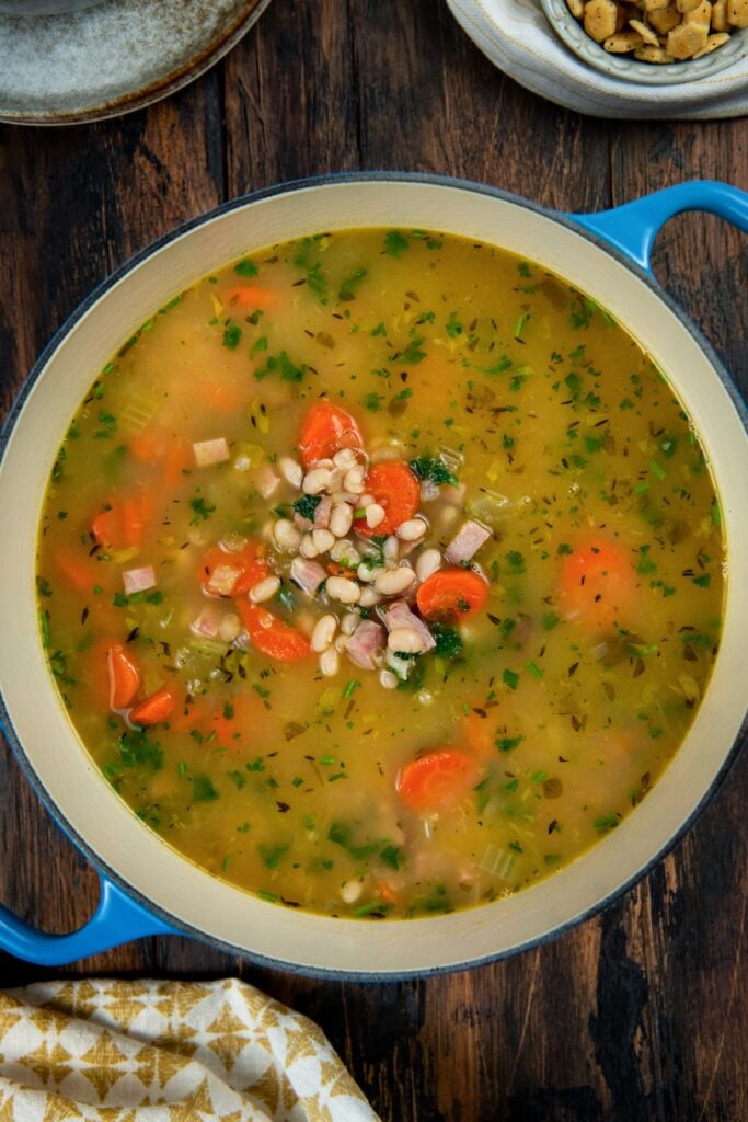 Made with dried or canned beans, this hearty Navy Bean Soup recipe is packed with creamy white beans, smoky ham, and flavorful veggies. A pot of hearty soup filled with navy beans, chopped carrots, diced ham, and green herbs, sitting on a dark wooden table next to a bowl of crackers and a patterned napkin.