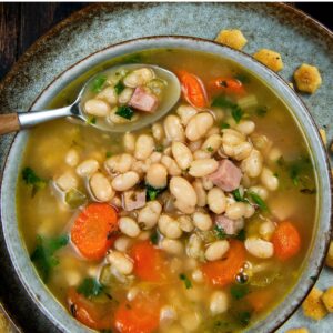 A bowl of navy bean soup with ham, carrots, celery, and parsley, served with oyster crackers on a rustic plate.