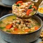 A close-up of a bowl of navy bean soup with ham, carrots, and greens. A spoon is lifting a serving of the hearty soup above the bowl.