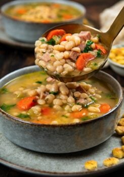 A close-up of a bowl of navy bean soup with ham, carrots, and greens. A spoon is lifting a serving of the hearty soup above the bowl.
