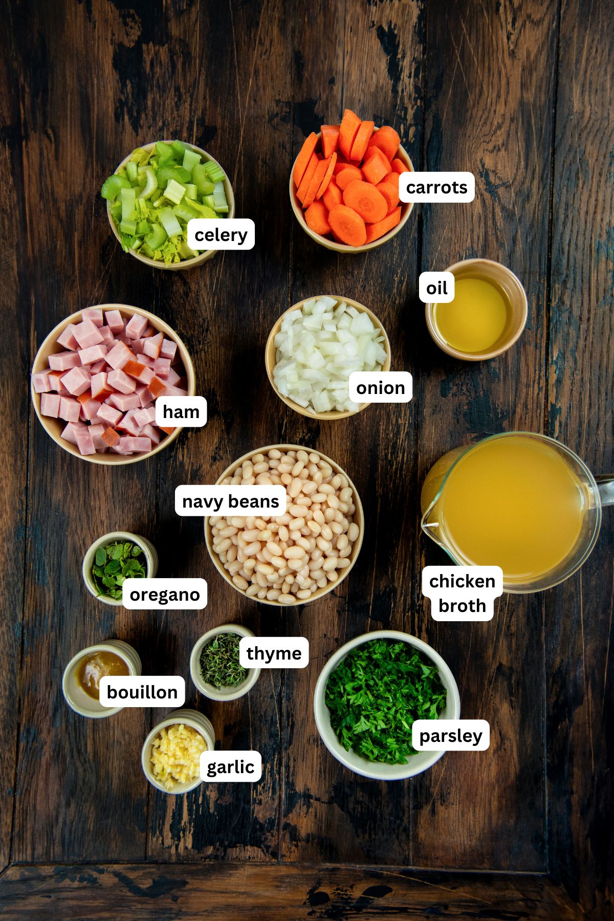 Made with dried or canned beans, this hearty Navy Bean Soup recipe is packed with creamy white beans, smoky ham, and flavorful veggies. Ingredients for navy bean soup recipe on a wooden table, each labeled: chopped celery, sliced carrots, oil, diced ham, chopped onion, chicken broth, navy beans, oregano, thyme, bouillon, minced garlic, and chopped parsley.