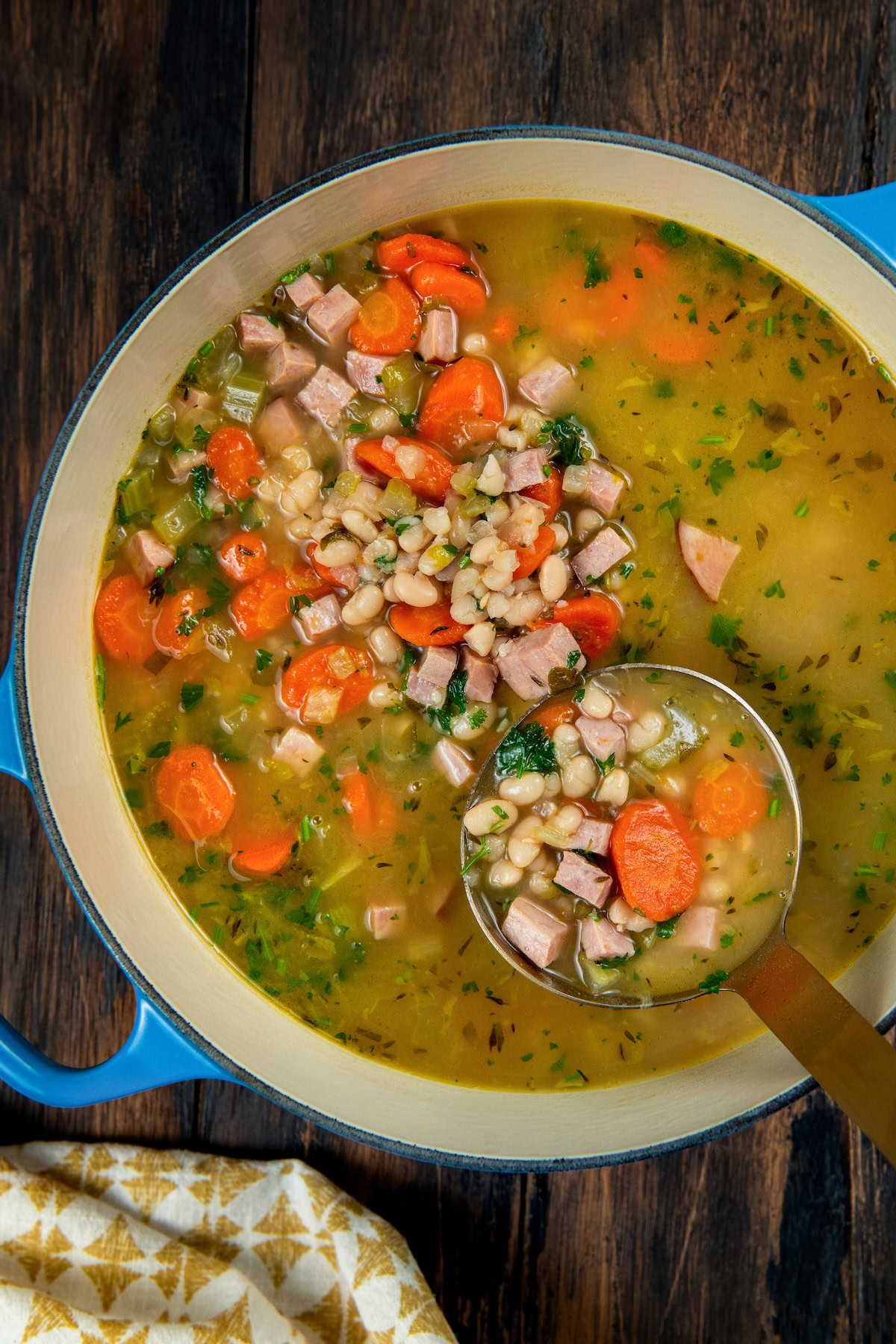 Made with dried or canned beans, this hearty Navy Bean Soup recipe is packed with creamy white beans, smoky ham, and flavorful veggies. A pot of navy bean and ham soup filled with sliced carrots, diced ham, white beans, and chopped herbs. A ladle is lifting some of the soup.