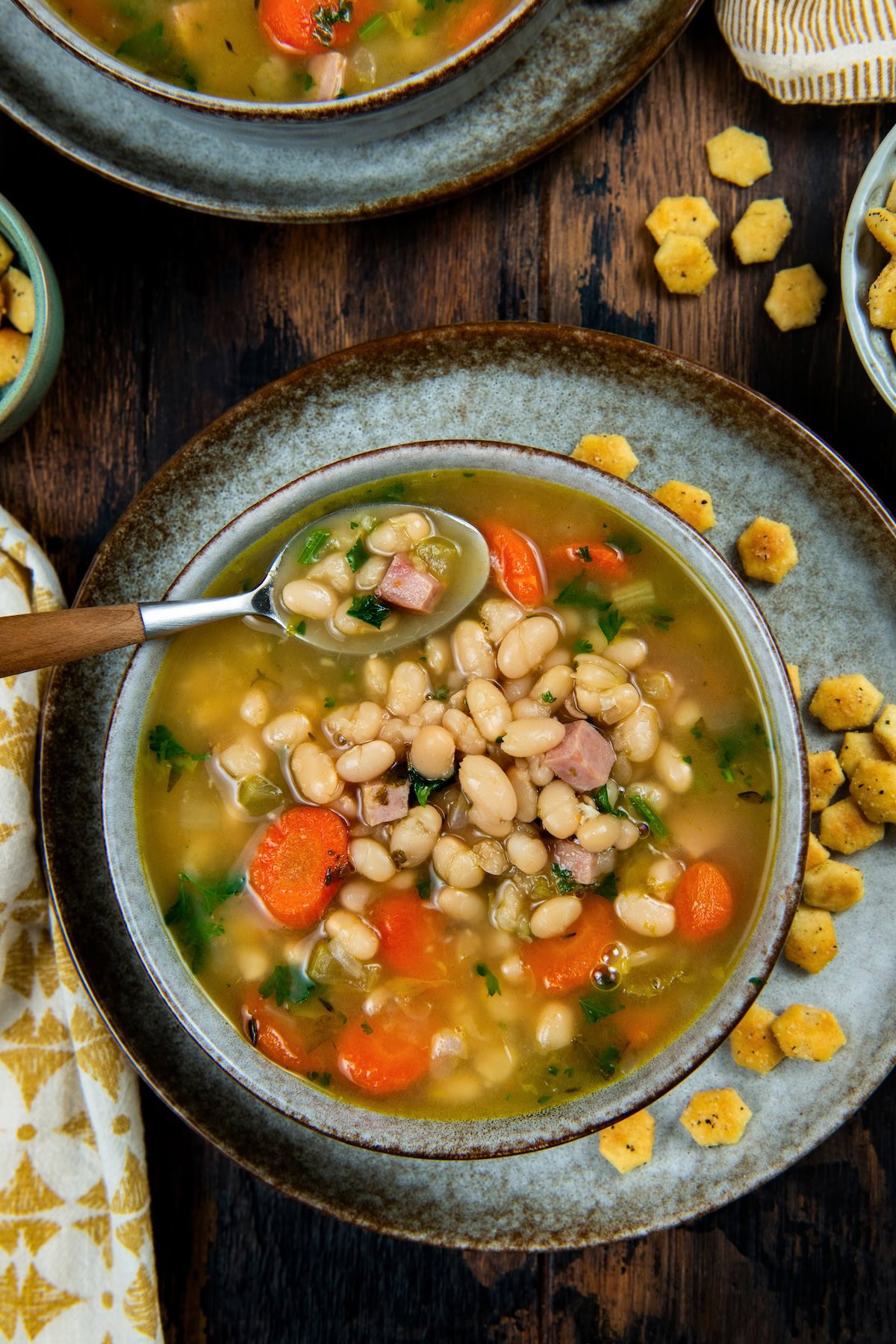 Made with dried or canned beans, this hearty Navy Bean Soup recipe is packed with creamy white beans, smoky ham, and flavorful veggies. A bowl of navy bean soup with carrots, celery, ham, and parsley, served with a spoon on a rustic plate.