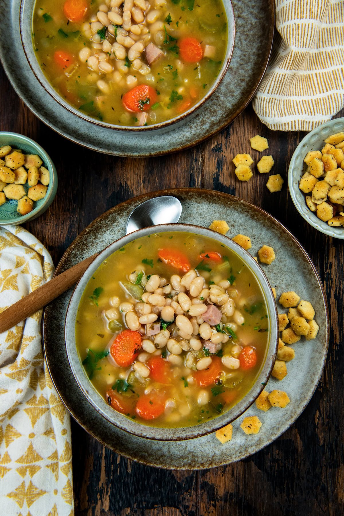 Made with dried or canned beans, this hearty Navy Bean Soup recipe is packed with creamy white beans, smoky ham, and flavorful veggies. Two bowls of navy bean soup with ham, carrots, and herbs on rustic plates, surrounded by small bowls of oyster crackers. A spoon rests on one of the bowls.