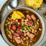 A bowl of black eyed peas with ham and chopped green onions, served with a buttered cornbread muffin on a plate, and a spoon on the side.
