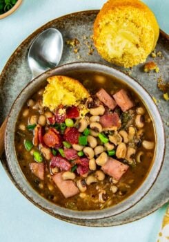 A bowl of black eyed peas with ham and chopped green onions, served with a buttered cornbread muffin on a plate, and a spoon on the side.