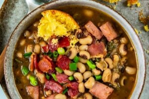A bowl of black eyed peas with ham and chopped green onions, served with a buttered cornbread muffin on a plate, and a spoon on the side.