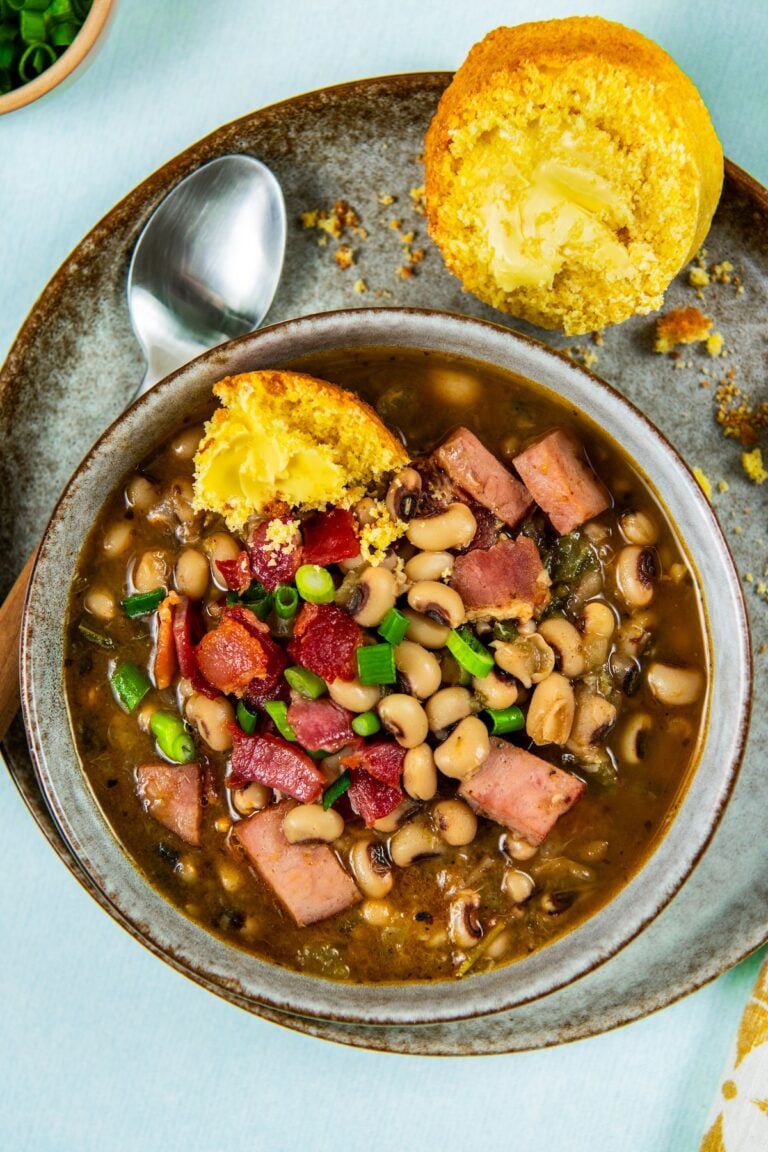 A bowl of black eyed peas with ham and chopped green onions, served with a buttered cornbread muffin on a plate, and a spoon on the side.